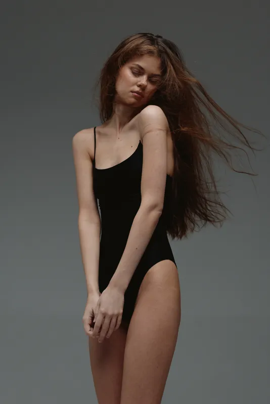 Young woman in black bodysuit posing with flowing hair against grey background, elegant, minimalistic, fashion studio shot