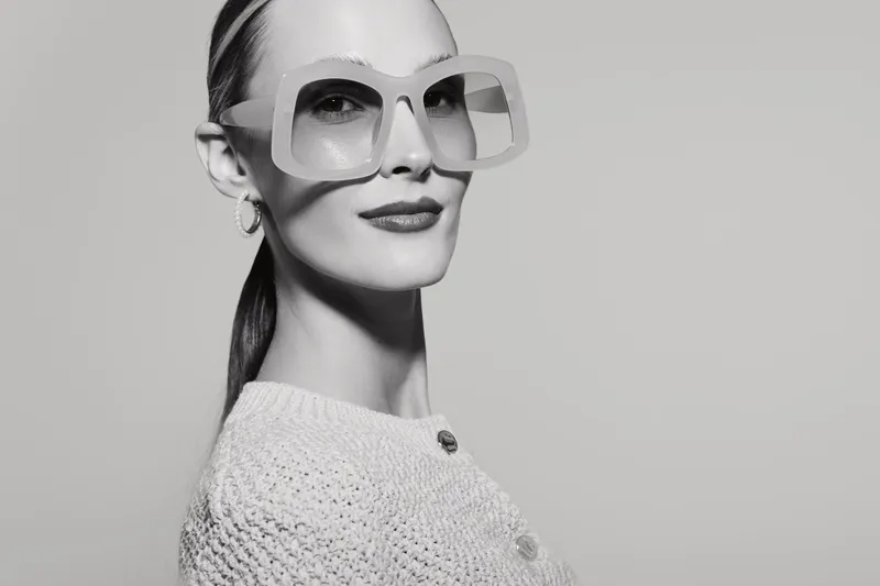 Stylish white woman with fair skin wears large square sunglasses and knitted sweater posing with confident smile on grey background.