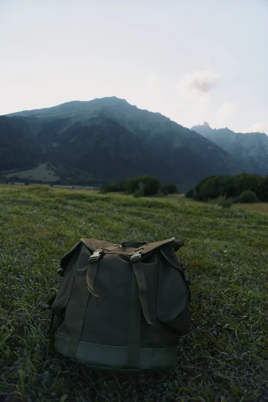 Rugged backpack resting in a grassy field with distant mountains, an outdoor lifestyle scene that conveys adventure, solitude, and simple travel gear in natural light