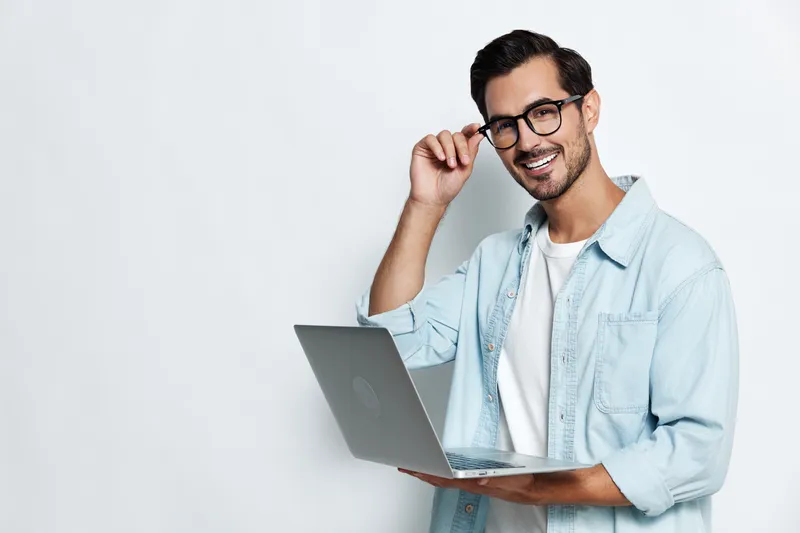 Stylish smiling young man wearing glasses and a denim jacket holds a laptop while looking at the camera against a light background. Professional lifestyle concept.
