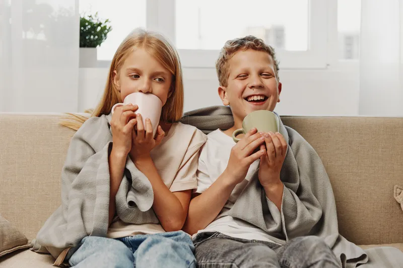 Two children wrapped in cozy blankets laugh and enjoy warm drinks sitting on a beige sofa in a bright living room.