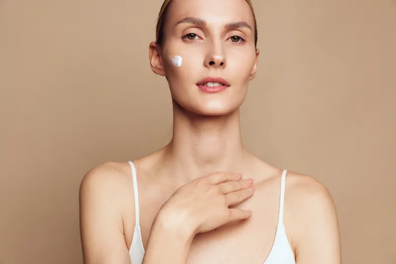 A young woman with clear skin applies moisturizer cream on her face and chest against a beige background. Concept for skincare, beauty, and self care routine.