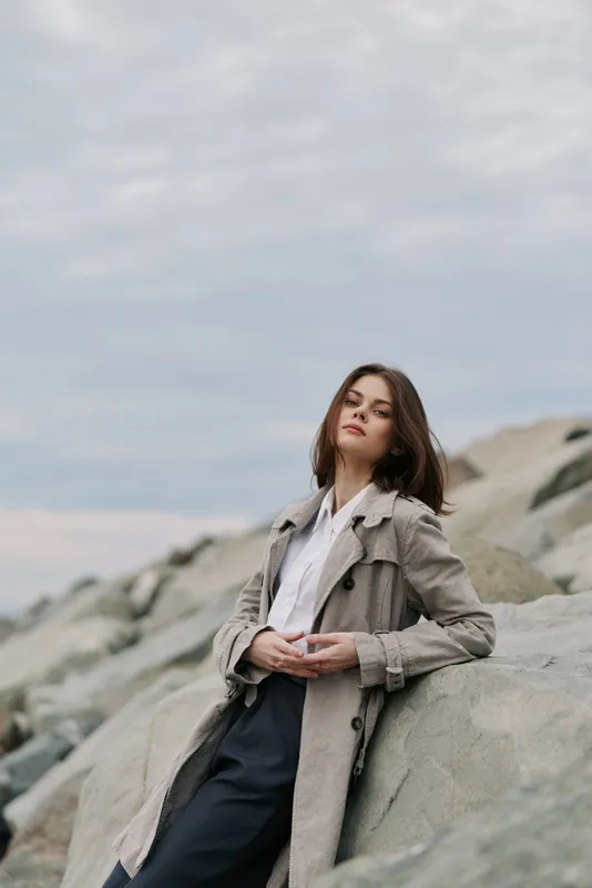 Young woman in stylish coat leans on large rocks outdoors under cloudy sky. Casual fashion portrait in natural light with relaxed pose and calm mood near rocky shore landscape.