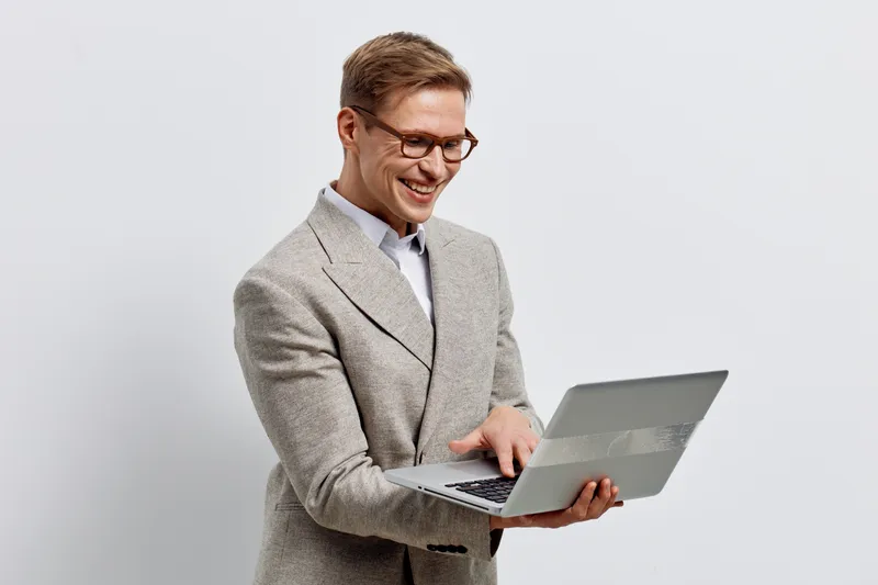Smiling young man with glasses in stylish gray blazer holding a laptop on plain light gray background. People business casual portrait, professional, modern lifestyle, technology, indoor. Concept: wor