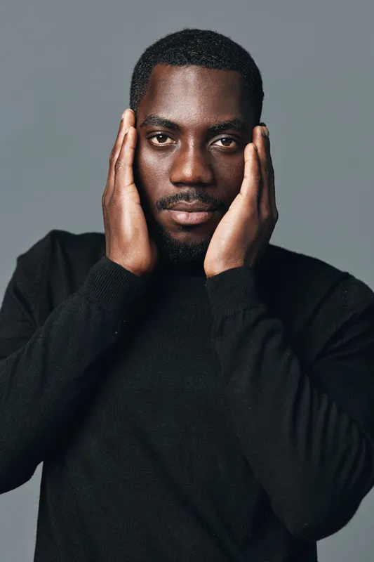 African man with dark skin and beard wearing black sweater posing with hands on face in creative conceptual visual style. Studio portrait with serious expression and direct eye contact.