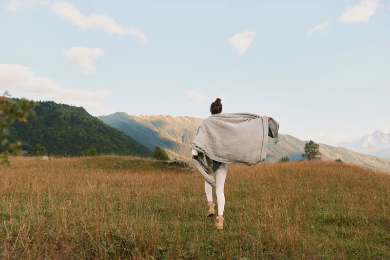 Woman running through a sunlit field with a flowing shawl, open landscape, outdoor lifestyle, freedom and exploration, bright sky, distant hills, calm mood, natural light, candid moment.