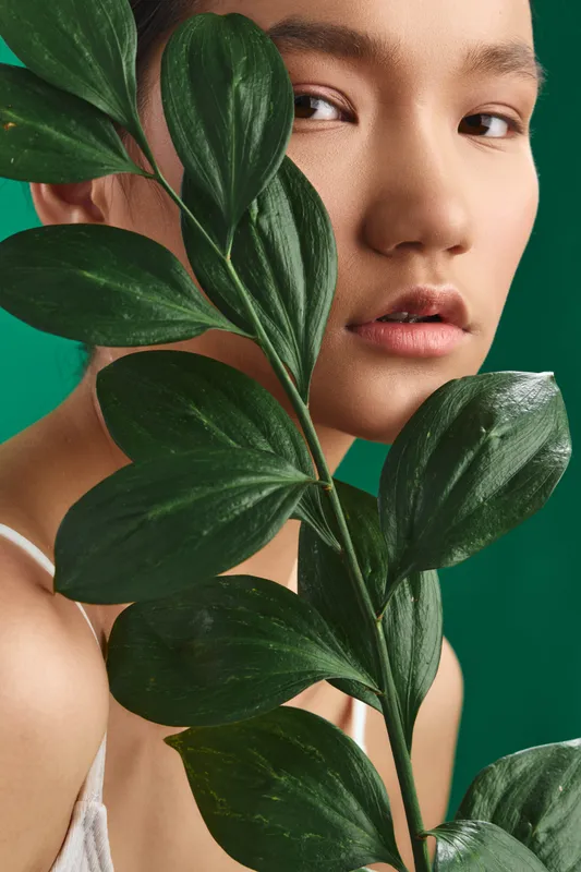Asian woman is photographed in a studio close up, partially hidden by lush leaves. She gazes softly at the camera, conveying calm, natural beauty and a fresh minimal aesthetic.