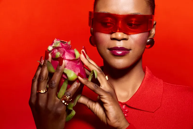 Close-up of African woman with dark skin holding dragon fruit. She wears red sunglasses and red outfit against red background. Concept of exotic fruit, fashion, and vibrant colors.
