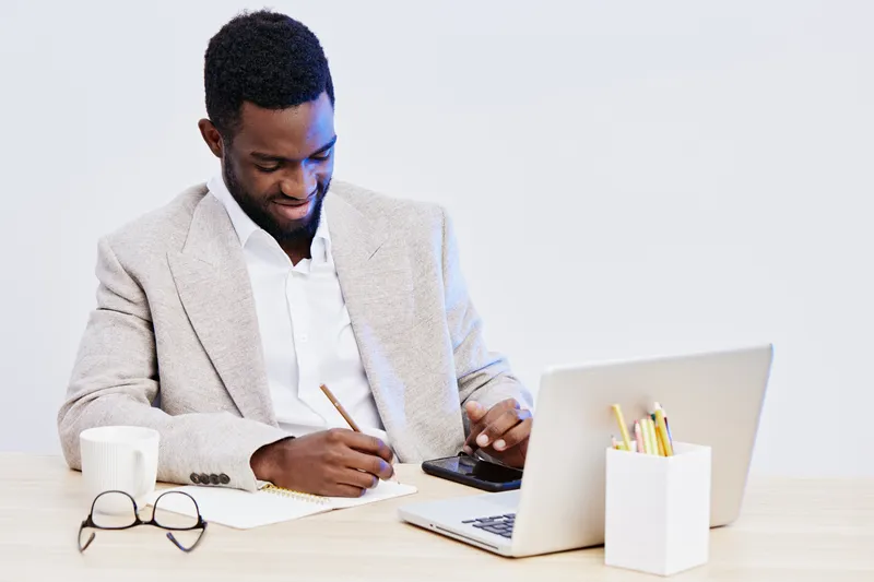 Professional black man in beige blazer and white shirt working at office desk, writing in notebook with laptop, glasses, coffee mug and supplies, modern workspace, business and productivity concept.