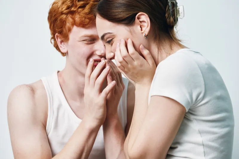 Young couple smiling closely with hands near faces in white casual clothes, showing affection and happiness in studio with neutral background.