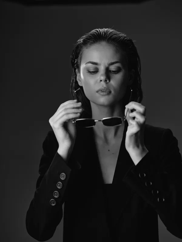 Black and white portrait of a serious woman holding sunglasses wearing a dark blazer under studio lighting.