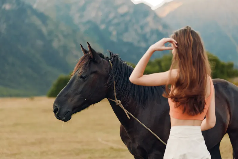 A woman stands beside a dark horse in an open field with distant mountains. She smiles gently, touching the horse's mane, creating a calm, natural outdoor moment.
