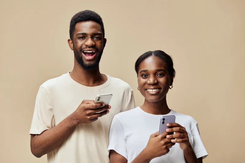 Young African couple happily using smartphones together, dressed in casual light colored clothing, smiling against a warm beige background