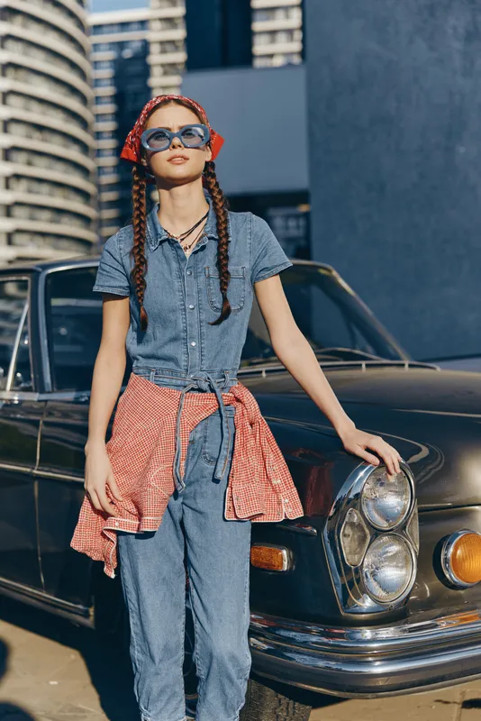 young woman with braids posing near vintage car wearing blue denim jumpsuit and red checkered shirt tied around waist under sunny clear sky