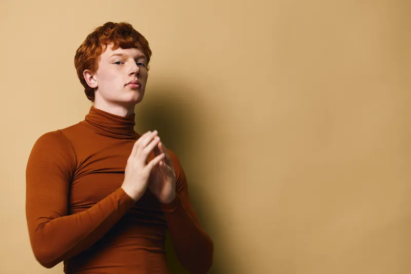 Young man with red hair wearing brown turtleneck sweater standing against beige wall with serious expression and hands clasped in front of chest in thoughtful pose.