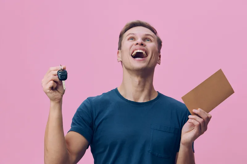 Enthusiastic young man smiling happily, holding a set of keys in one hand and a cardboard envelope in the other, wearing a casual blue t-shirt, celebrating success on a pink studio background. People 