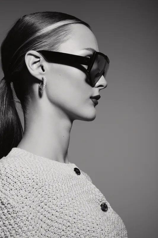 Profile of a young white woman wearing large sunglasses and a knitted sweater, posing in a studio with soft lighting. Fashion and style concept.