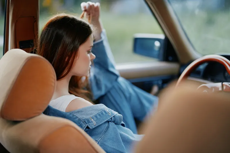 Young woman in denim outfit sitting in a vintage car, expressing relaxation and contemplation, with a vibrant natural background and soft focus details.