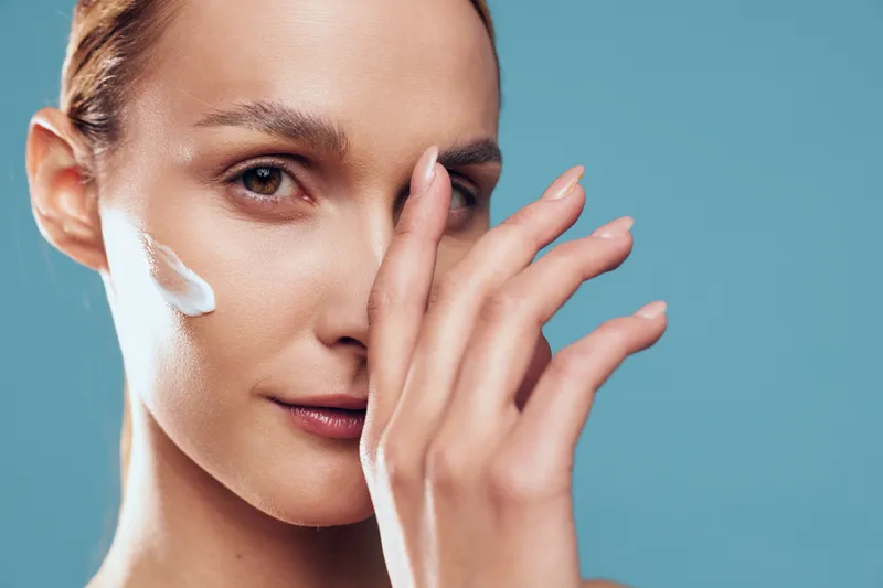 Closeup portrait of woman applying moisturizing cream on face with fingers, skincare routine, clear skin, natural makeup, soft lighting, blue background, calm expression
