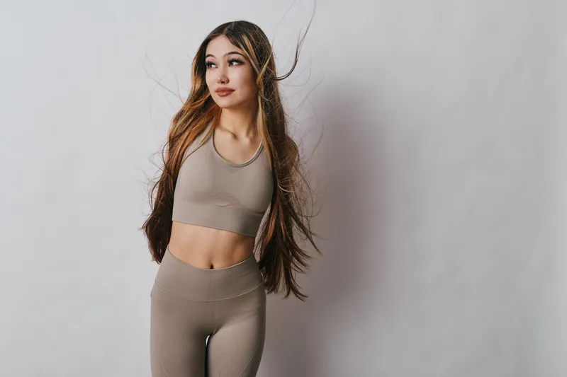 Young woman in activewear posing with loose long hair and confident expression for fitness, wellness and lifestyle portrait on light background indoors.