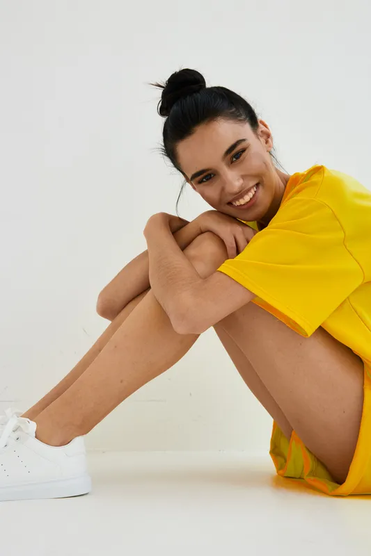 smiling woman in yellow shirt sitting with folded arms around bent knees, casual fashion, white sneakers, natural light, studio background, joyful mood