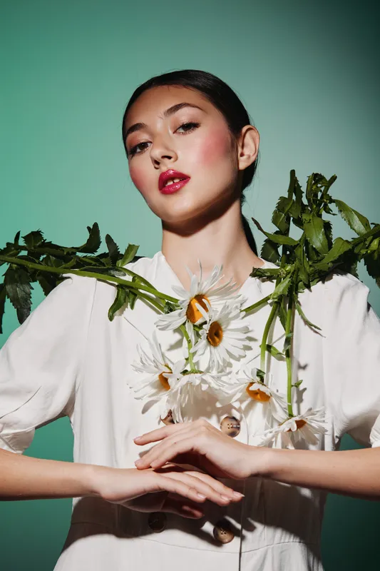 Elegant woman wearing white outfit poses with daisies and green stems as fashion accessories against turquoise background in a creative beauty concept.