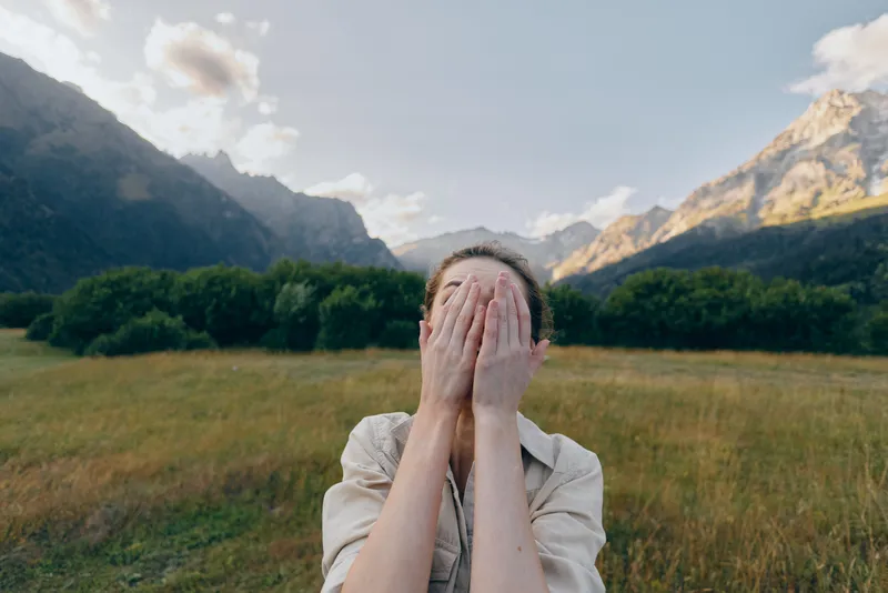 Woman outdoors in a grassy field with mountains in the background, hands covering her face, conveying playful surprise and a candid lifestyle moment.
