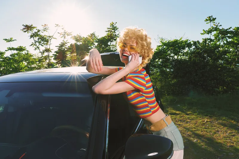 Cheerful woman outdoors, leaning on a car, laughing with eyes closed, sunlit moment, striped shirt, breezy atmosphere, retro colors, natural backdrop