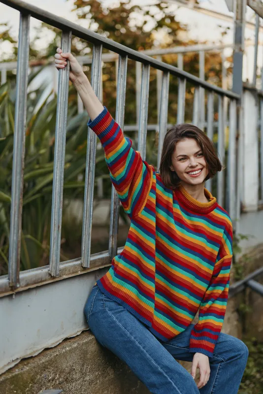 Woman smile sweater rainbow sitting by a metal railing outdoors in casual jeans, relaxed pose and happy portrait with cozy knit, natural light and urban garden background