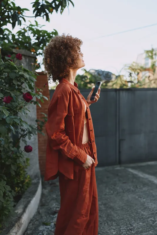 Woman lifestyle fashion outdoor orange suit curly hair holding smartphone, urban setting, confident mood, natural light, casual elegance, everyday activity, modern vibe, candid moment