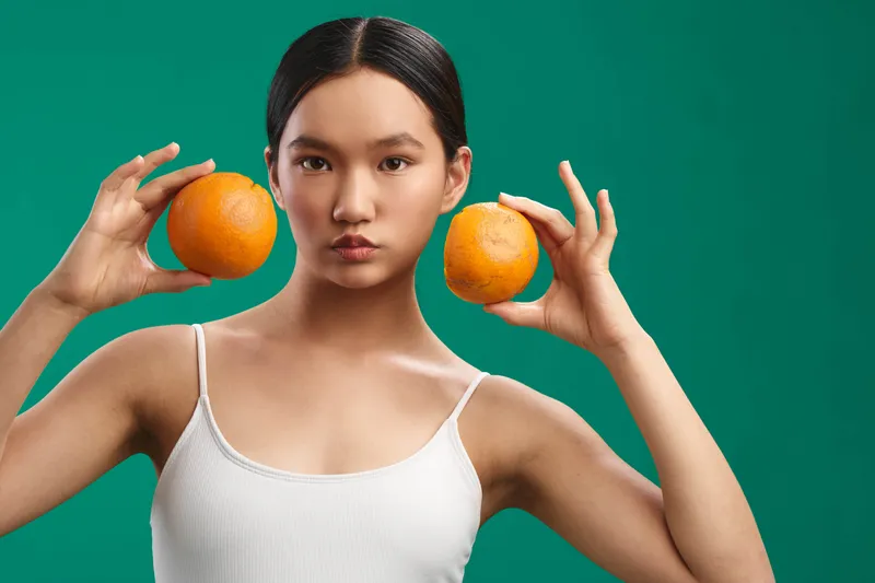 Asian woman holds oranges in a bright studio, wearing a white top. The portrait captures a fresh, playful mood with bold color and balanced lighting.