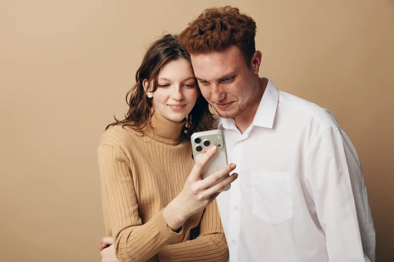 Young couple with casual clothes looking at smartphone screen together, smiling and sharing moment. Neutral beige background creates warm and cozy atmosphere of connection and technology.