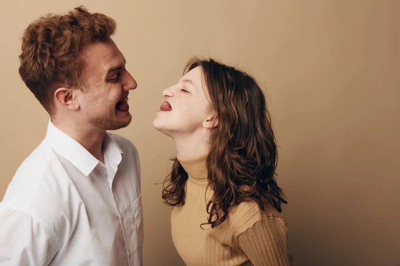 Young couple playfully sticking out tongues at each other, smiling and enjoying a lighthearted moment. Casual clothing and neutral background create a warm, fun, and intimate atmosphere.