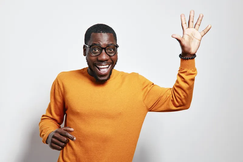African American man with dark skin wearing glasses and orange sweater smiling and waving hand in studio. Happy male portrait with casual clothing and positive gesture on white background.