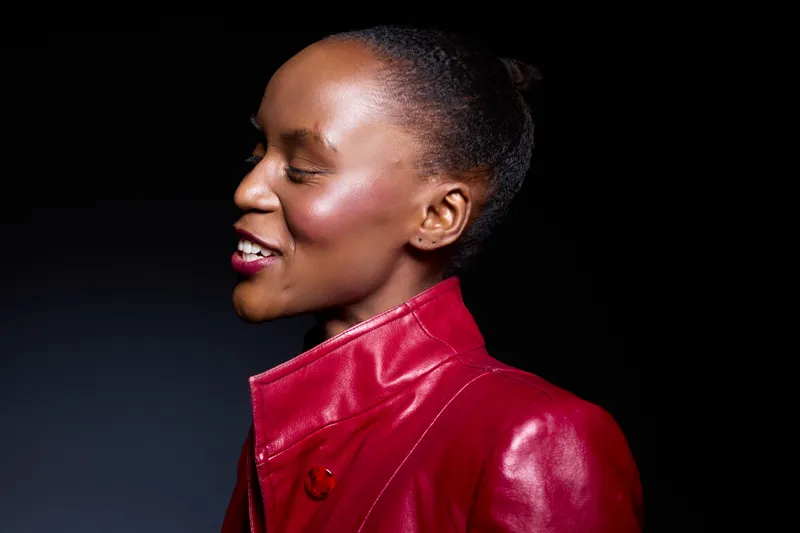 Profile of a smiling African woman with dark skin wearing a red leather jacket. Studio portrait with black background. Concept of fashion, confidence, and natural beauty in women.