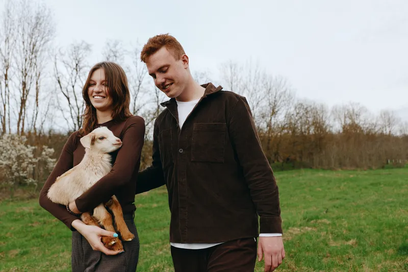 Joyful couple outdoors with a goat kid, walking through a green field, warm light, smiling faces, relaxed posture, natural tones, carefree moment, countryside vibe, gentle breeze vibrant