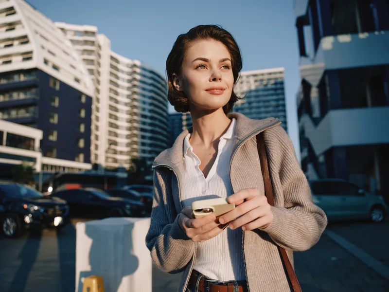 A confident businesswoman walks along an urban street, glancing at her smartphone with a calm smile, projecting focus, optimism, and professional clarity in a modern setting.