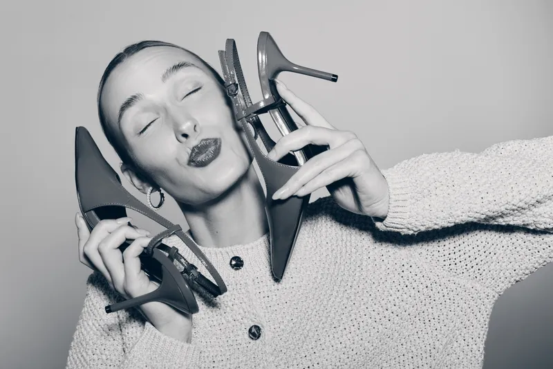 Young white woman with pale skin holds two high heel shoes near face with closed eyes and puckered lips. Black and white studio portrait with fashion and style concept.