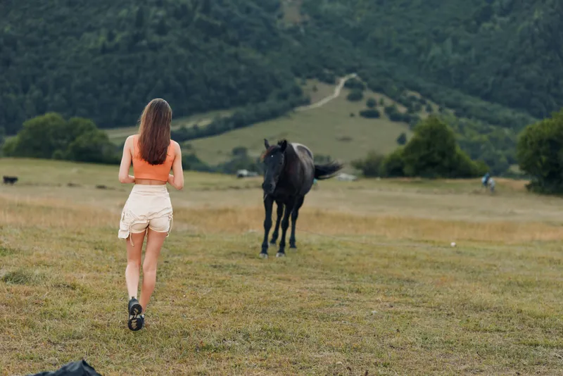 A young woman runs across a grassy field toward a horse under distant rolling hills, capturing a moment of nature, freedom, curiosity, and outdoor exploration in a serene countryside setting.