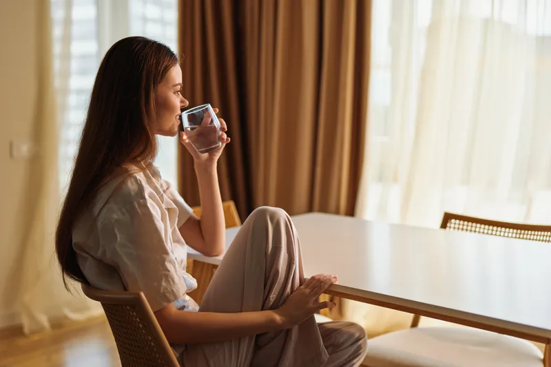 woman drink glass home morning at table, sitting relaxed in profile by window with soft sunlight and curtains. Casual female portrait enjoying hydration and a peaceful domestic routine.