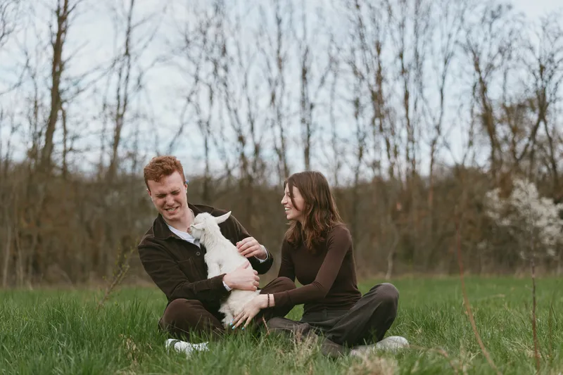 Couple in a grassy park smiles at a playful goat kid as they cuddle and share a moment of outdoor joy, highlighting lifestyle, affection, and connection with nature.