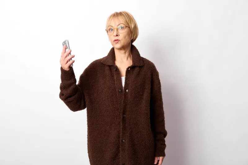 Surprised elderly woman holding a smartphone, wearing a brown coat in studio against white background. Technology communication portrait with curious expression, indoor fashion.