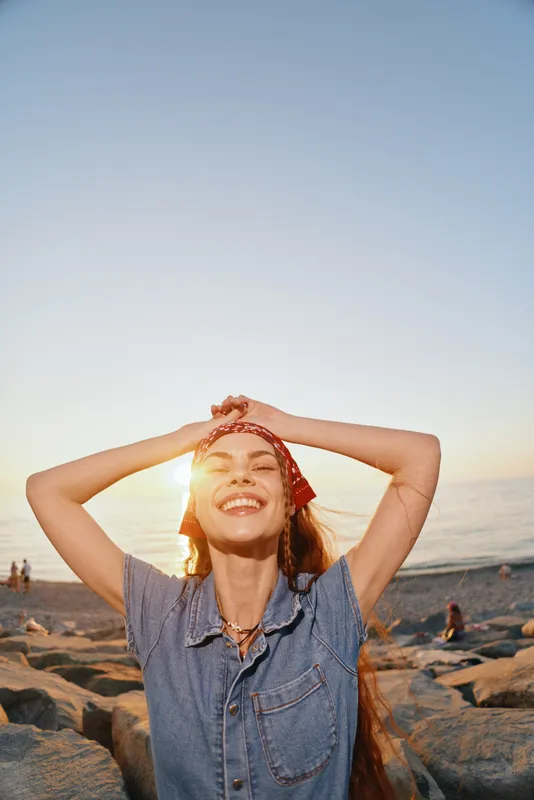 Lifestyle woman in modern boho-western denim street style enjoys sunny beach moment with joyful smile, film color tones enhance warm, carefree atmosphere.