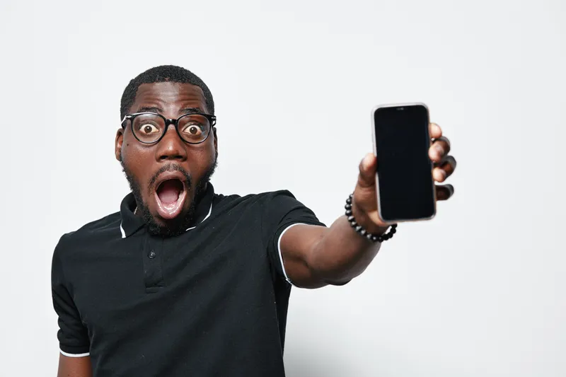 African American man with dark skin and glasses showing smartphone with surprised expression on white background. Black male holding phone closeup for technology and communication concept.