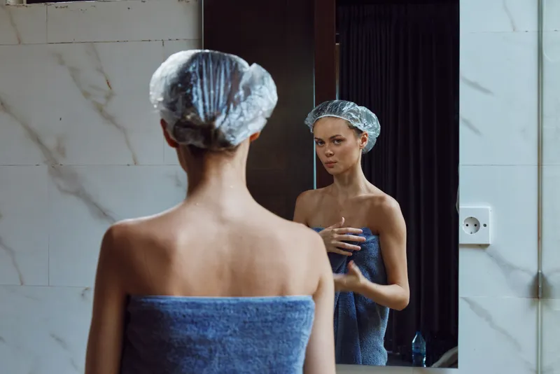 Woman in bathroom wearing shower cap and blue towel, looking at reflection in mirror, skincare routine, indoor, natural light, self-care moment.