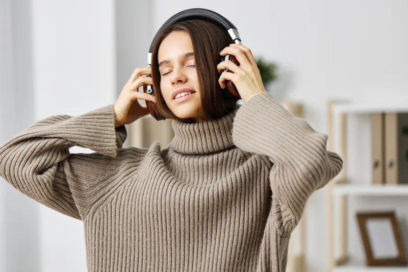 Молодая женщина европеоидной расы сидит в помещении в наушниках и улыбается, глядя в смартфон