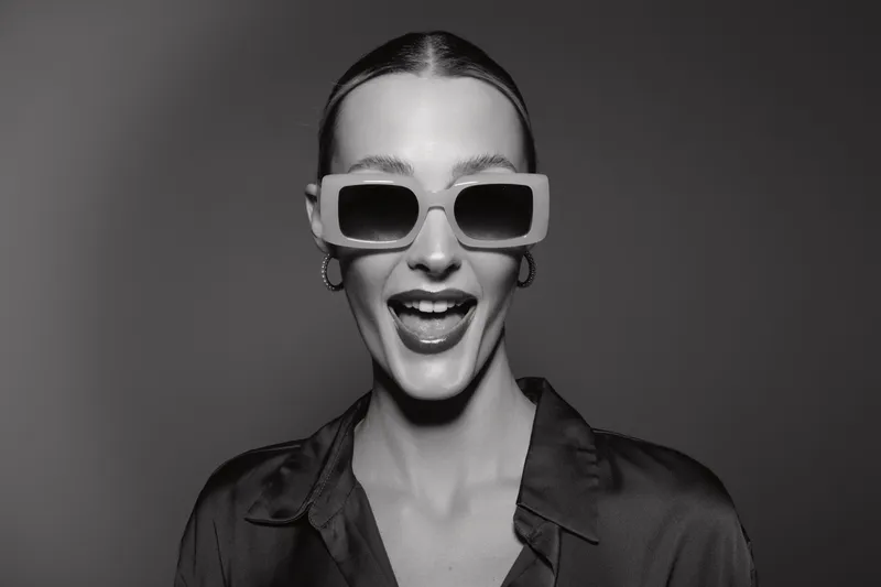 Happy white woman with pale skin smiling in oversized sunglasses and dark blouse on grey background. Concept of fashion, joy, and modern style.