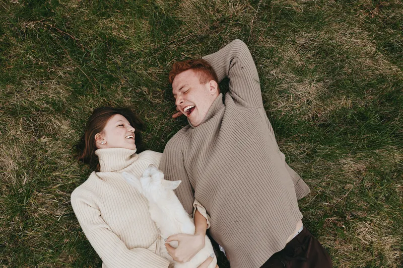 Young redhead man and brunette woman lie on grass laughing while holding a white baby goat, enjoying a cozy lifestyle moment outdoors in warm sweaters on a casual day.