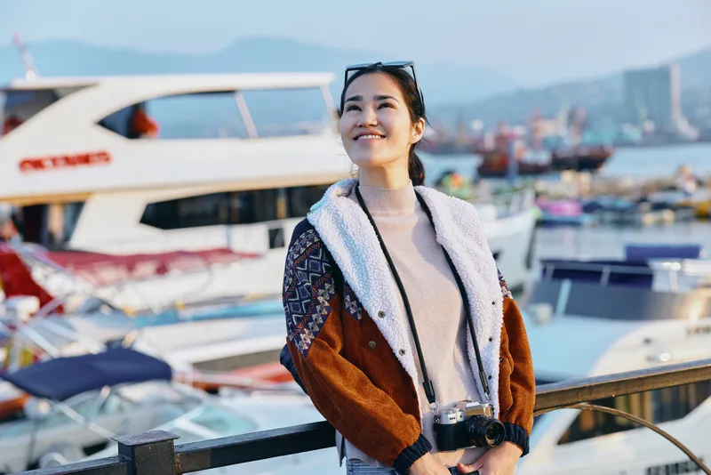 Woman in a cozy jacket with sunglasses by a busy marina, smiling, holding a drink, stylish travel vibe and outdoor leisure beside boats and shimmering water