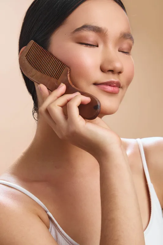 Asian woman is shown gently brushing her hair with a wooden comb, eyes closed and a soft smile, in a calm studio setting that conveys self care and wellness.